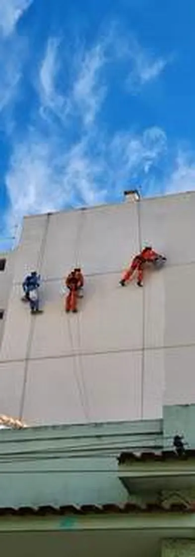 Três profissionais em alpinismo industrial, pendurados por cordas, trabalhando na fachada de um prédio